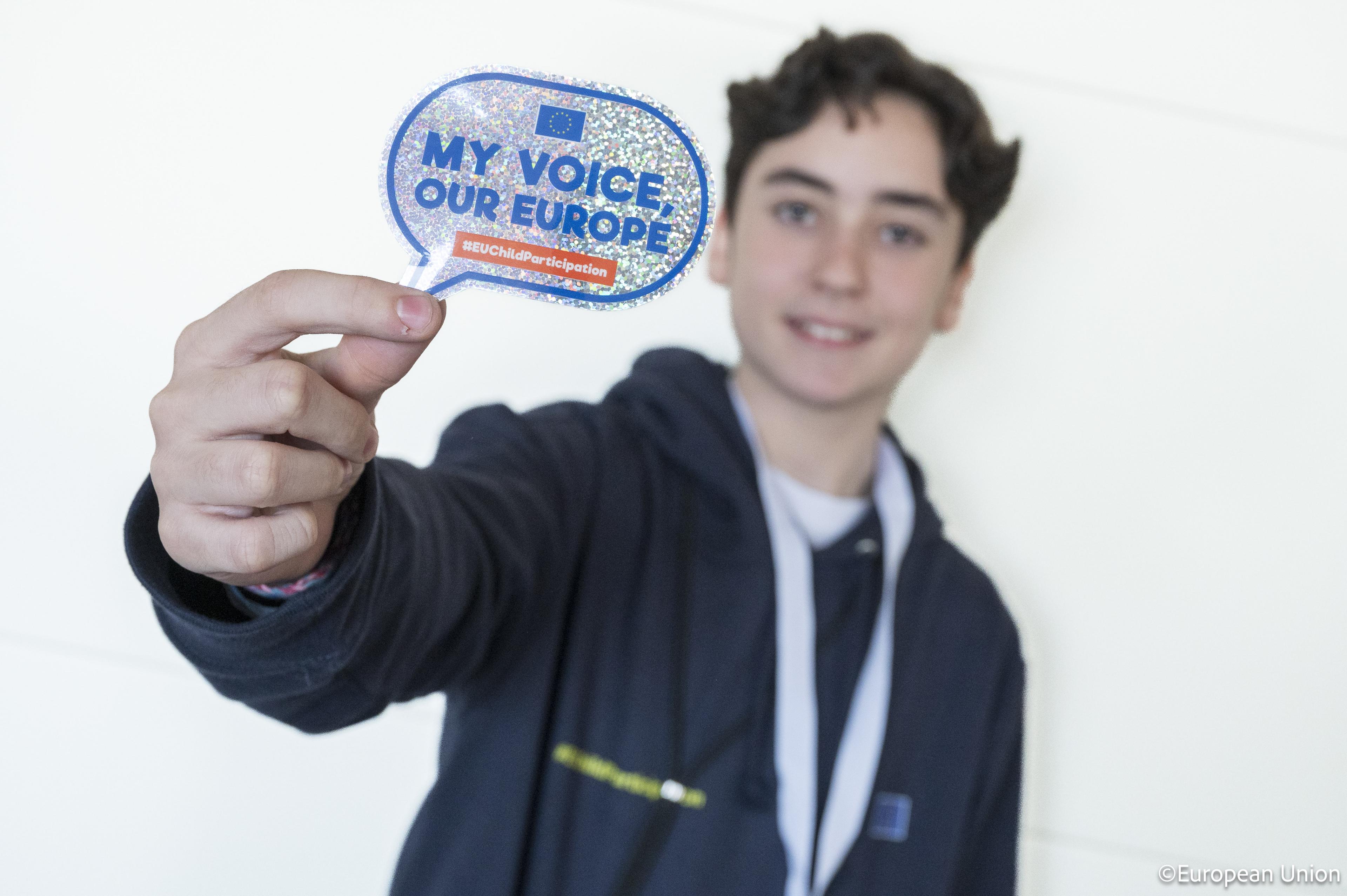 A young person holds up a colourful speech bubble sign that reads “My Voice, Our Europe” in front of a white background. The focus is on the sign, which promotes youth participation in Europe.
