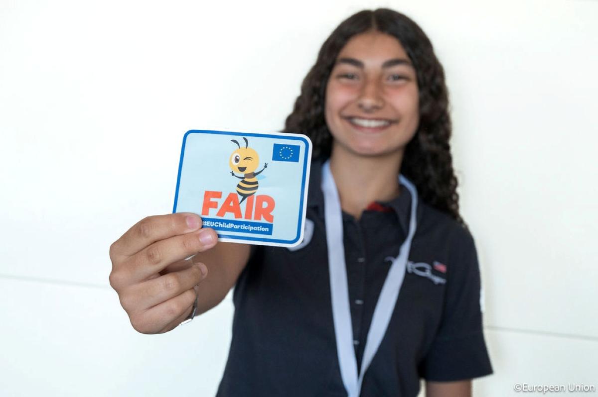 A young girl holding up a sticker with Be Fair on it.