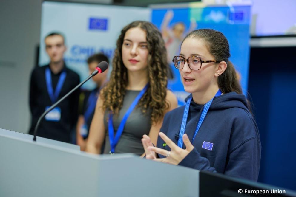 Two teenage girls speaking into a microphone, presenting their ideas to adults.